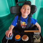 A boy in a wheelchair uses switches and joysticks on his lap tray.