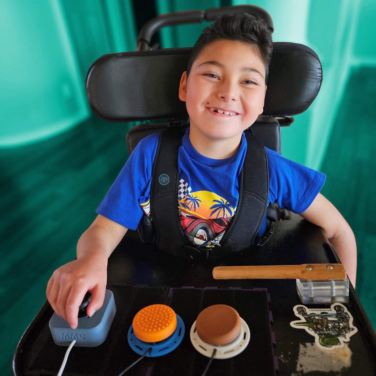 A boy in a wheelchair uses switches and joysticks on his lap tray.