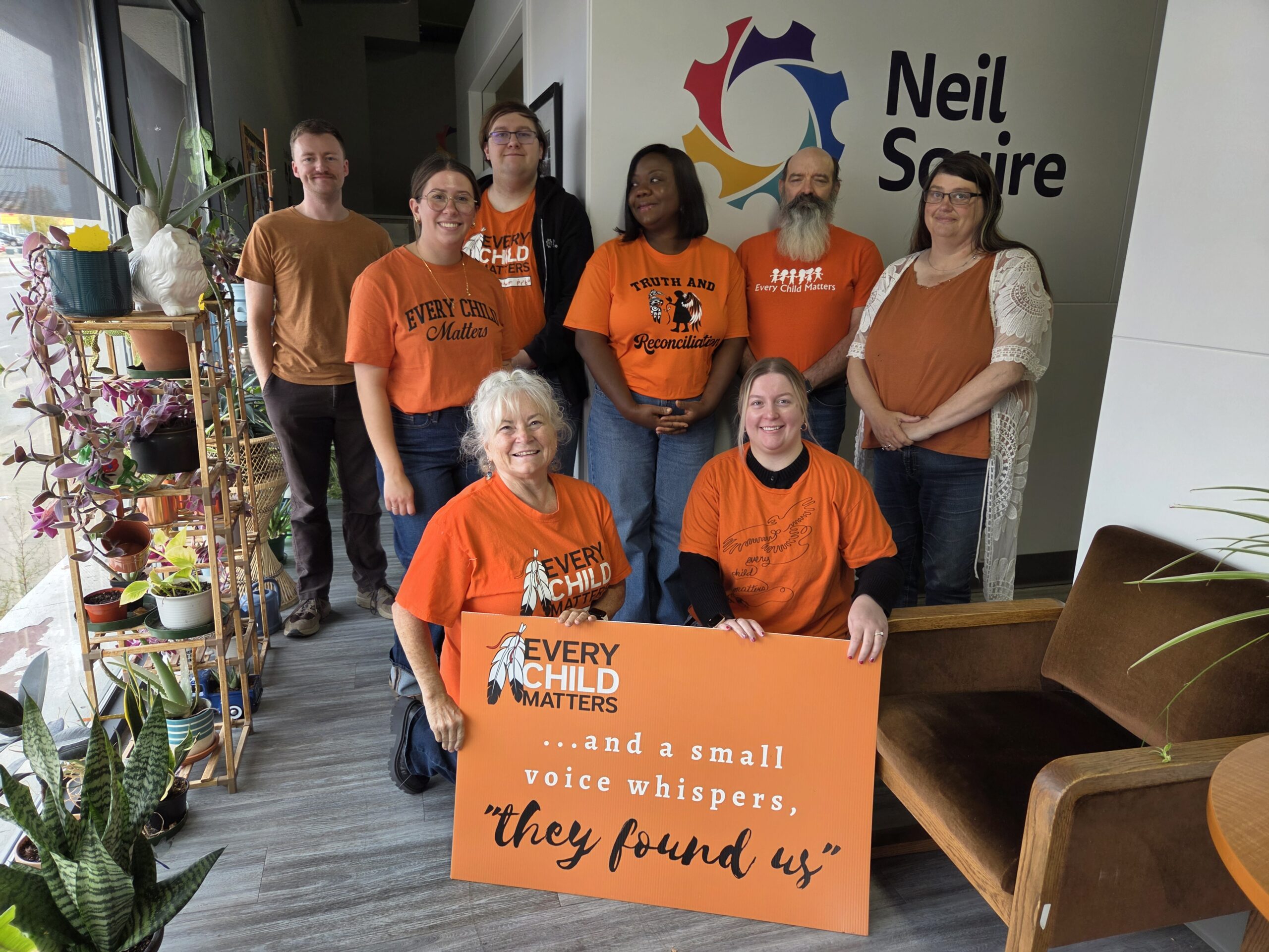 Our Regina office wearing orange shirts that say Every Child Matters and holding a banner that says, "...and a small voice whispers, they found us."