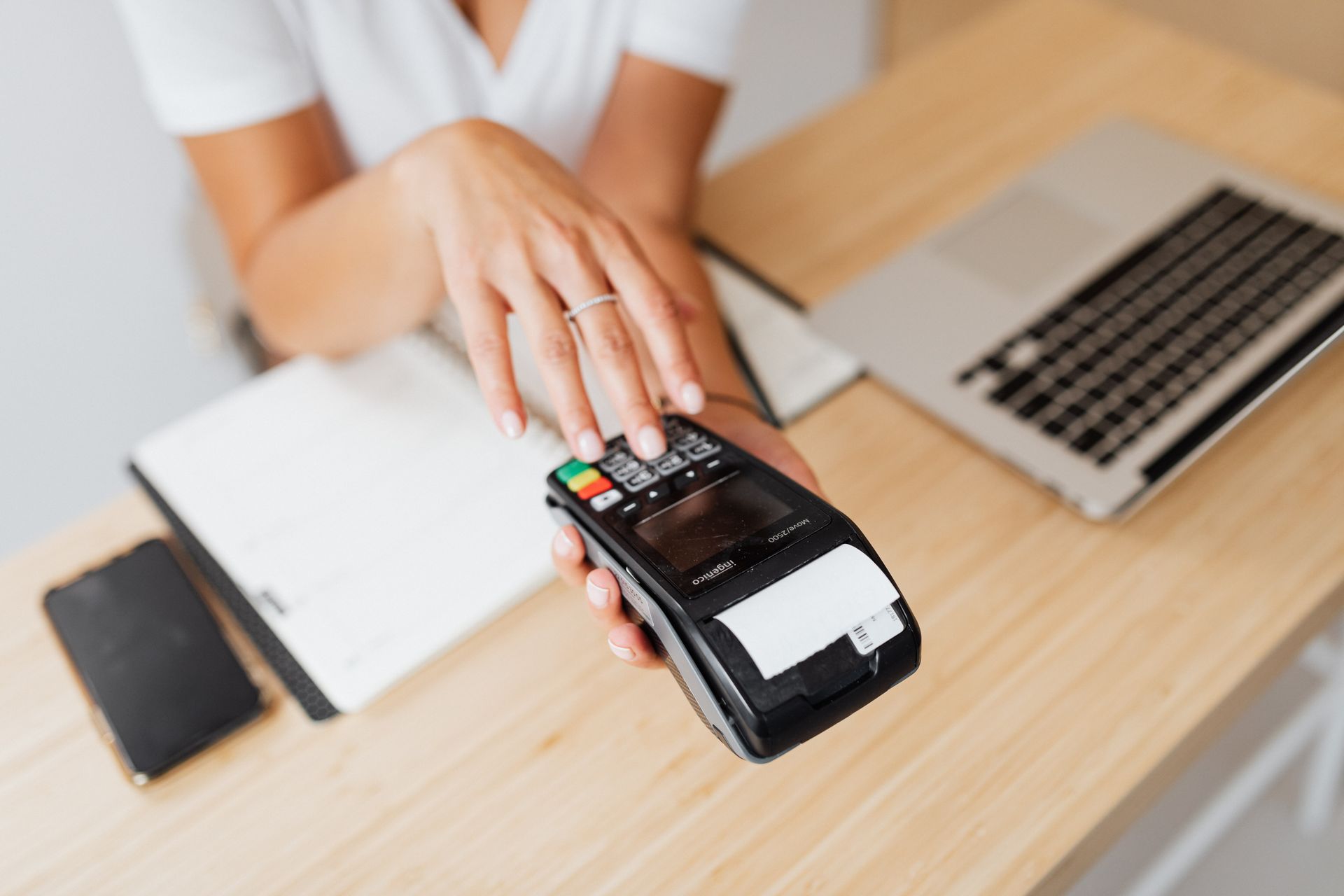 Person making a contactless payment by tapping a card on a portable card reader at a wooden desk with a laptop and notebook.
