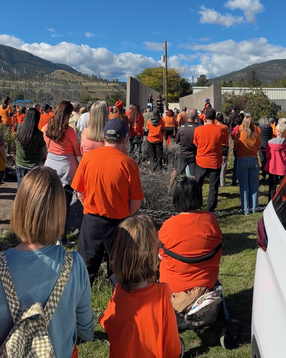 A large group of people wearing orange shirts gathered outdoors listening to a speaker at the Syilx Indian Residential School (SIRS) Monument.