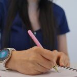 Person wearing a smartwatch writing notes with a pink pen in a spiral notebook on a wooden table.