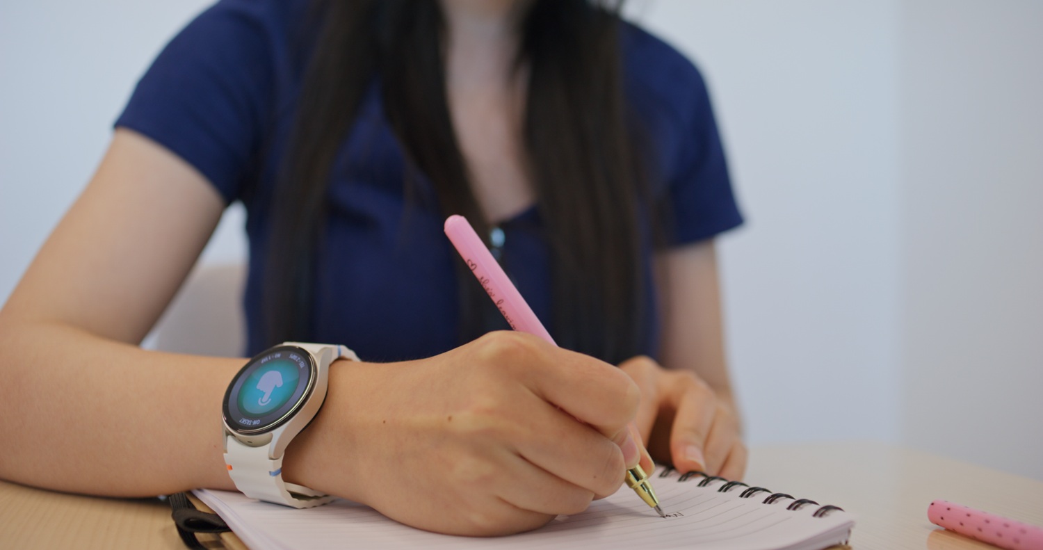 Person wearing a smartwatch writing notes with a pink pen in a spiral notebook on a wooden table.