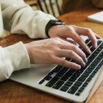 Person typing on a laptop keyboard at a wooden desk.