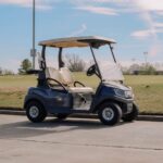 Golf cart parked on a paved path next to a grassy golf course under a partly cloudy sky.