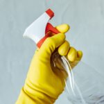 Hand wearing a yellow rubber glove gripping a clear spray bottle with a red and white nozzle against a plain background.