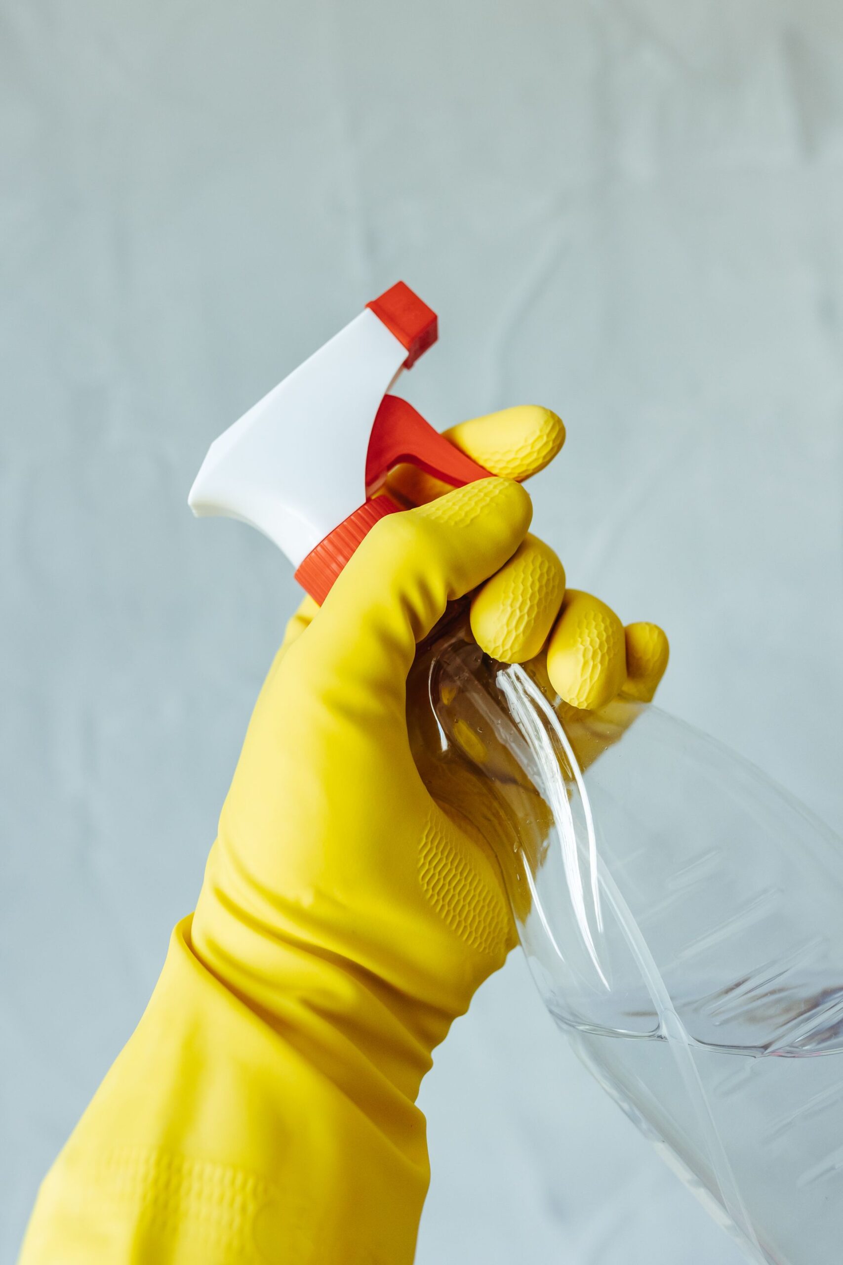 Hand wearing a yellow rubber glove gripping a clear spray bottle with a red and white nozzle against a plain background.